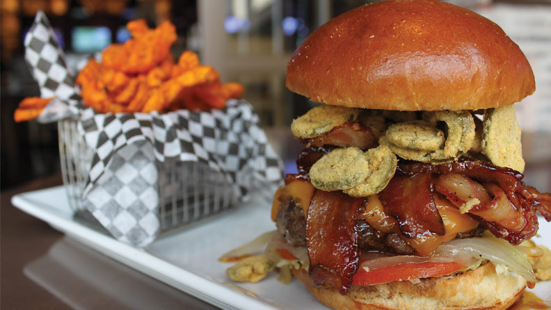 burger with bacon, cheese, tomato, onions and fried jalapenos on a plate with a container of sweet potato fries